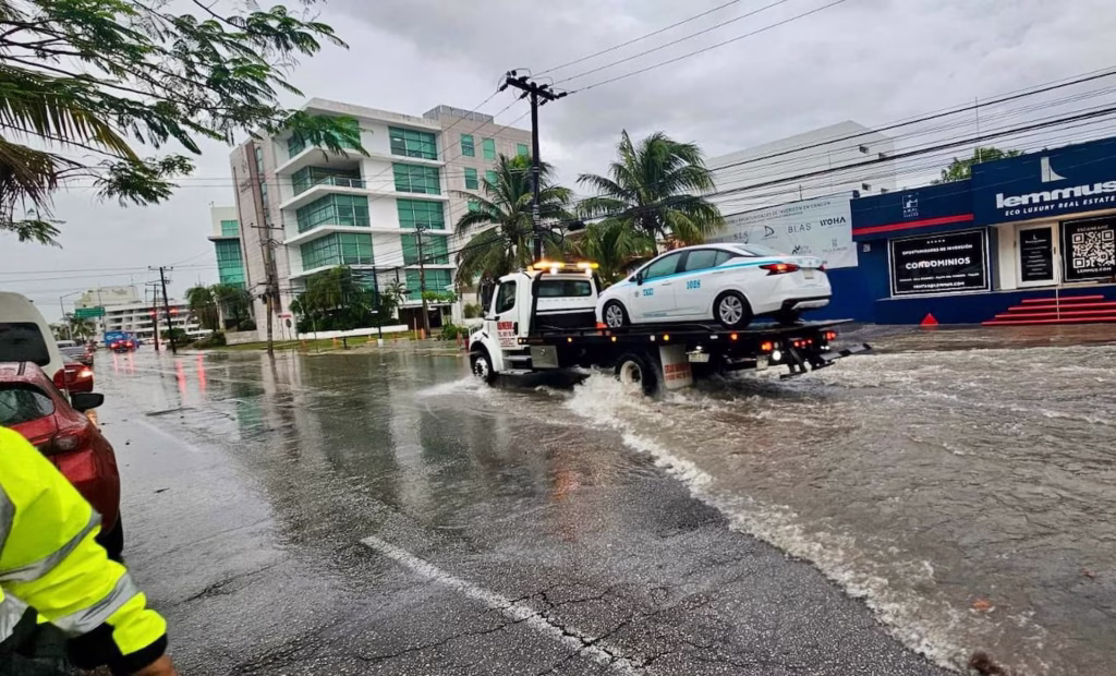 Grúa retira un vehículo afectado por inundaciones en una avenida de Cancún tras fuertes lluvias.