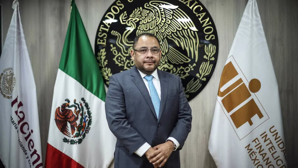 Funcionario de la Unidad de Inteligencia Financiera posa frente al escudo nacional y las banderas de México y de la UIF.