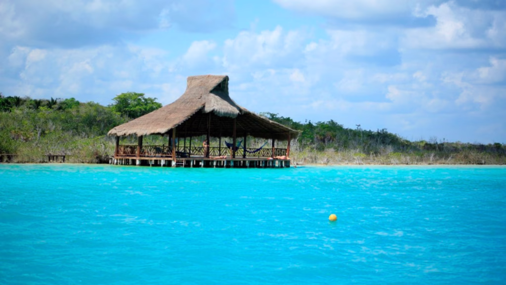Visitantes permanecen en un muelle con palapa frente a las aguas de la Laguna de Bacalar.