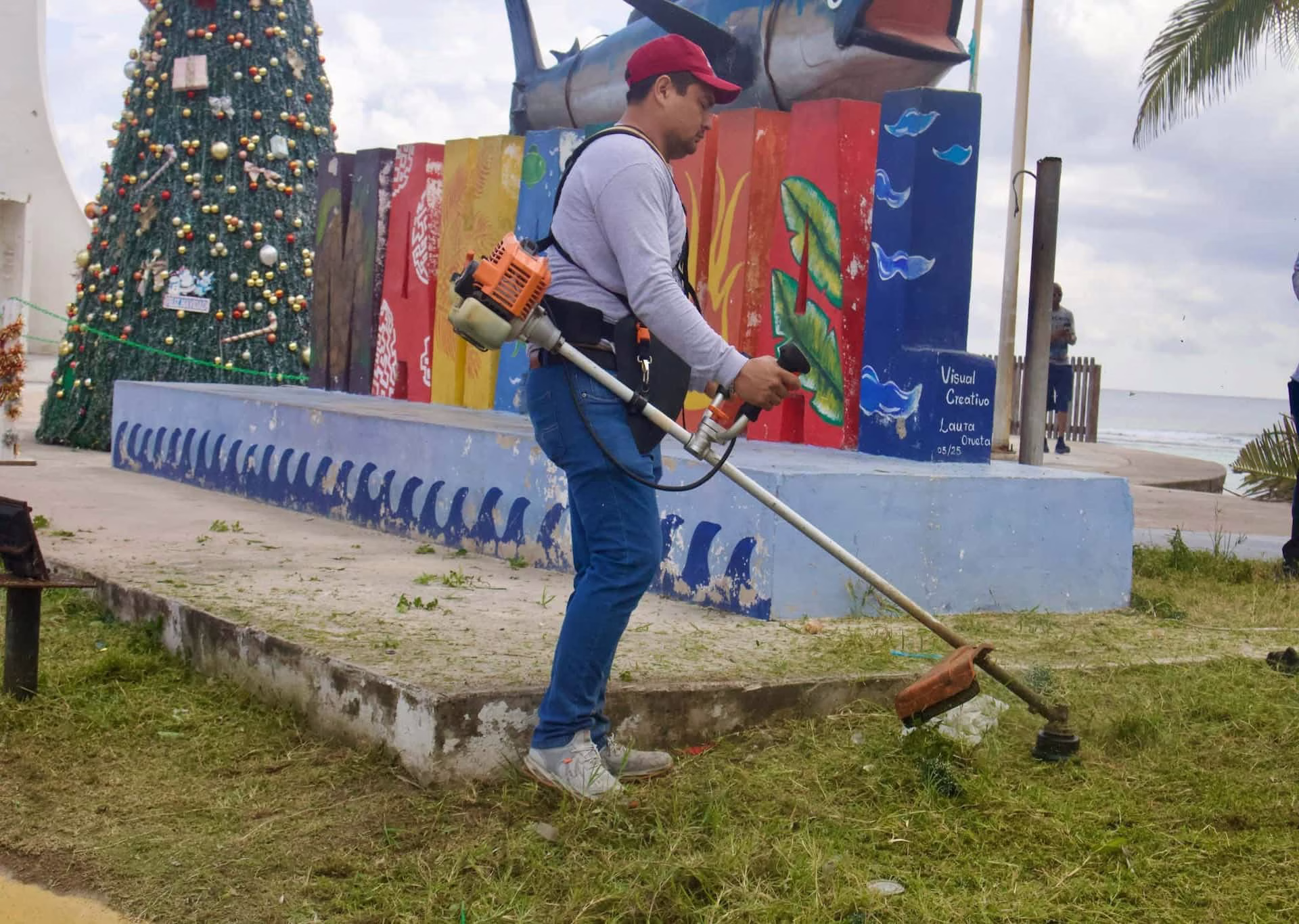 Trabajador realiza labores de mantenimiento en un área verde de Mahahual, donde el cuidado de los espacios públicos forma parte de la imagen turística del destino.