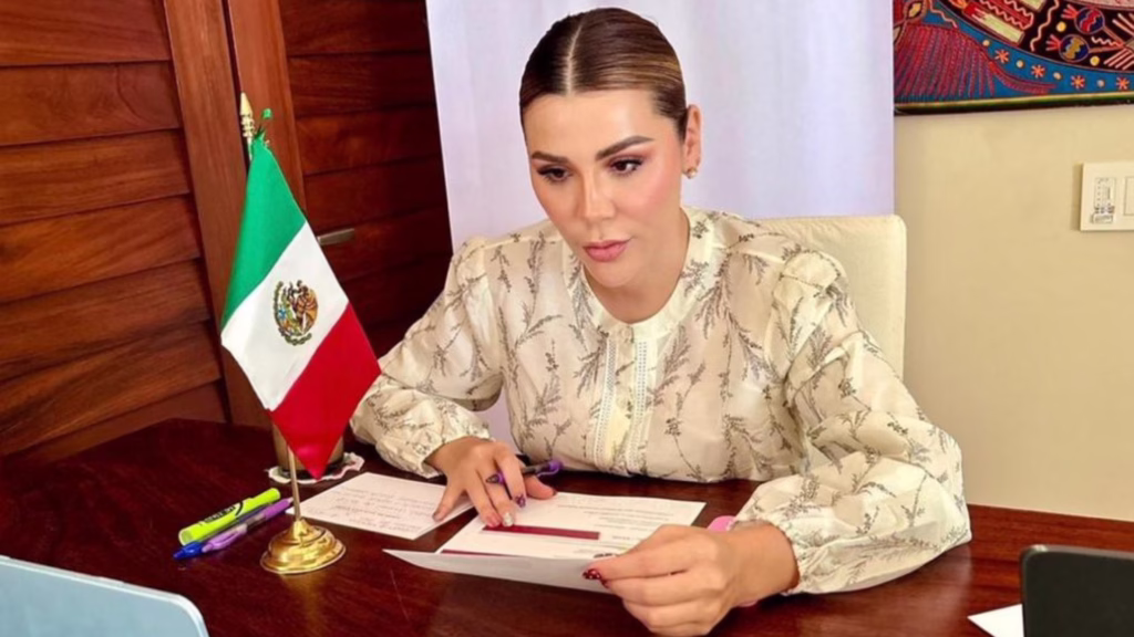 Marina del Pilar revisa documentos desde un escritorio, acompañada por una bandera de México.