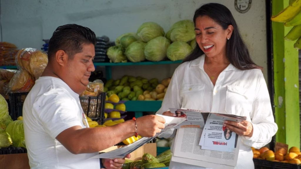Marybel Villegas conversa con un comerciante mientras ambos sostienen ejemplares de un periódico en un mercado de Cancún.