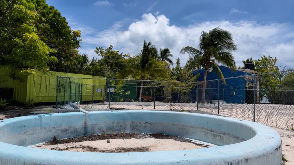 Área de la Tortugranja de Isla Mujeres con estructura circular vacía, cercado y construcciones al fondo.