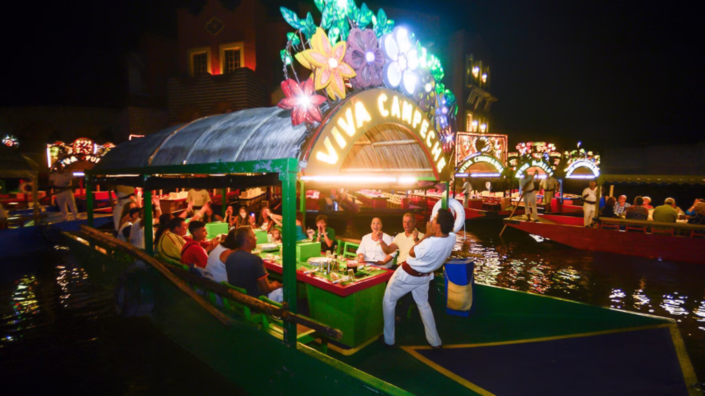 Visitantes disfrutan de una trajinera iluminada durante una experiencia nocturna de temática mexicana en Cancún.