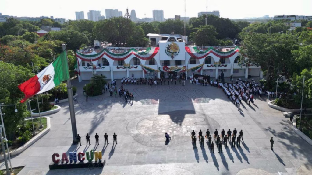 Vista aérea de la explanada del Palacio Municipal de Cancún con bandera monumental y formación cívica.