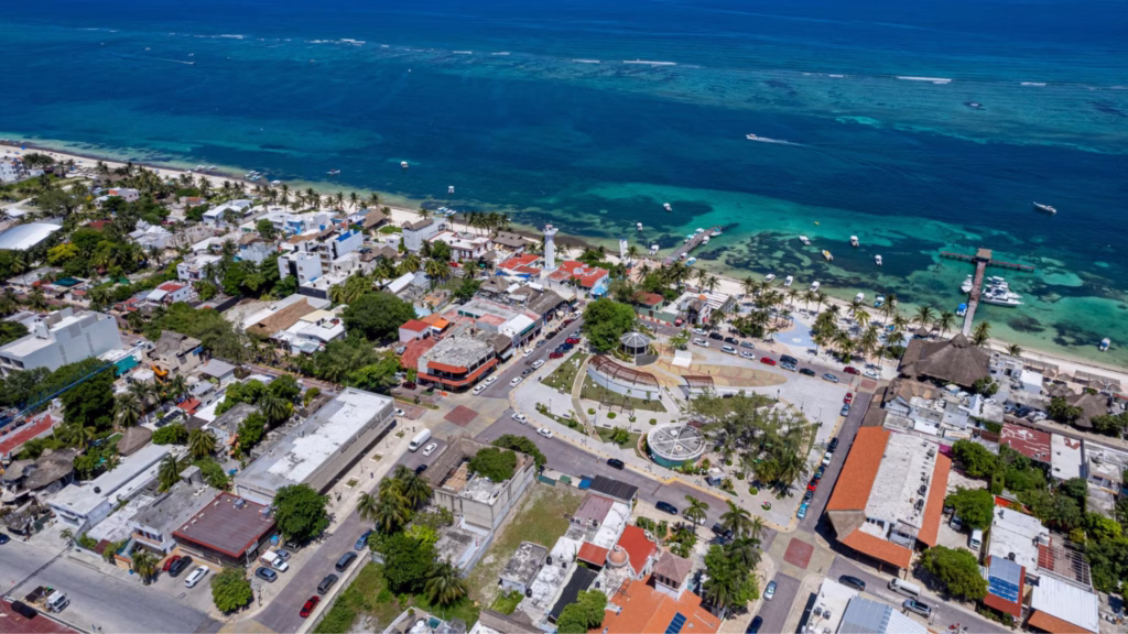 Vista aérea de una zona costera con playa, muelle, embarcaciones y zona urbana cercana al litoral. Se aprecia una comunidad turística del Caribe mexicano con infraestructura habitacional y comercial.